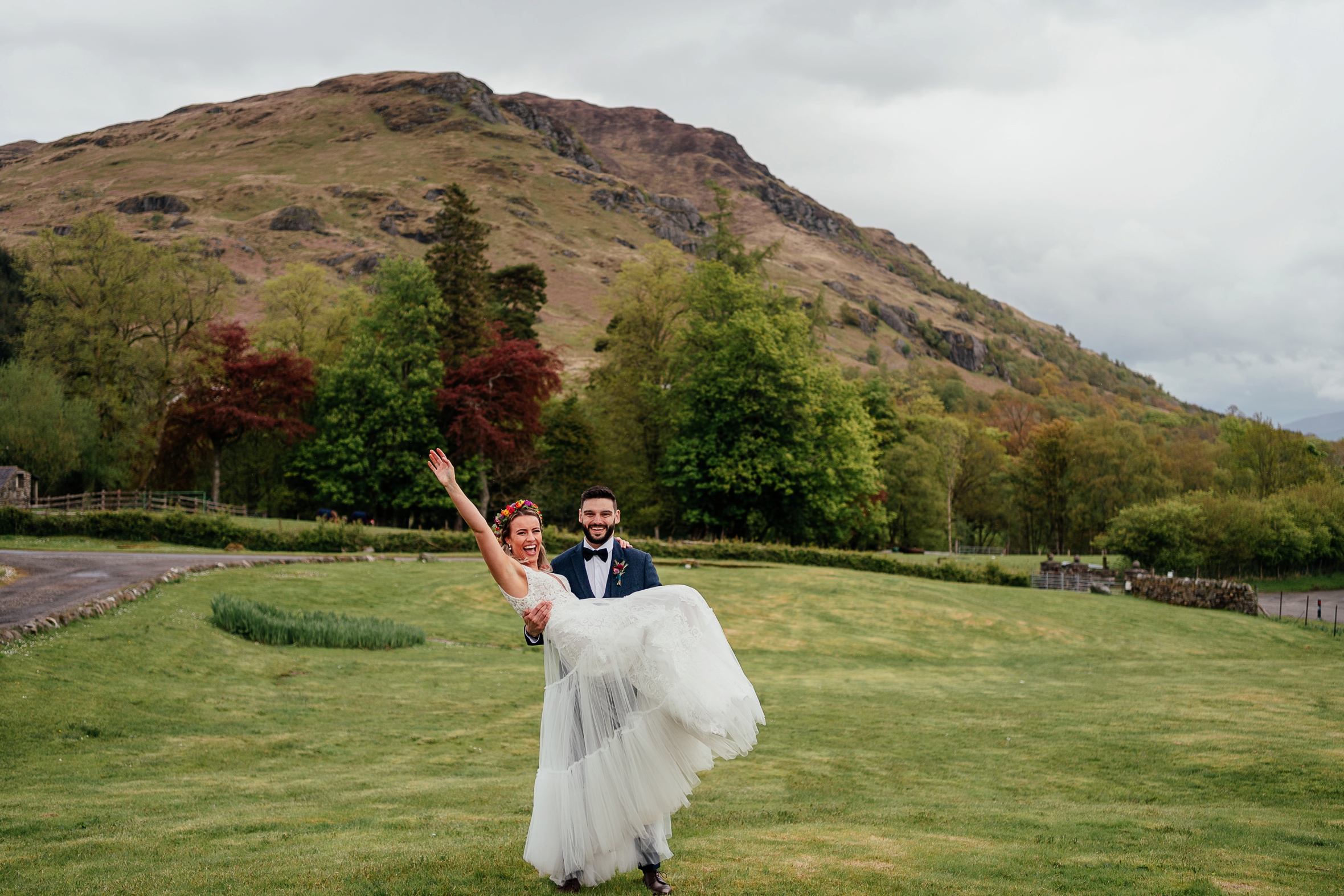 groom holding bride in arms on grass lawn bride wearing colourful flower headpiece monachyle mhor elopement