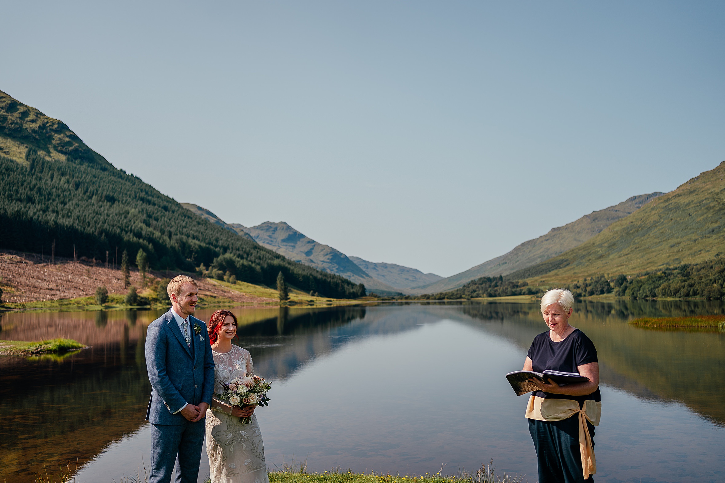 humanist celebrant reading to bride and groom at the tip of still loch with reflections of blue sky and green mountains humanist weddings scotland
