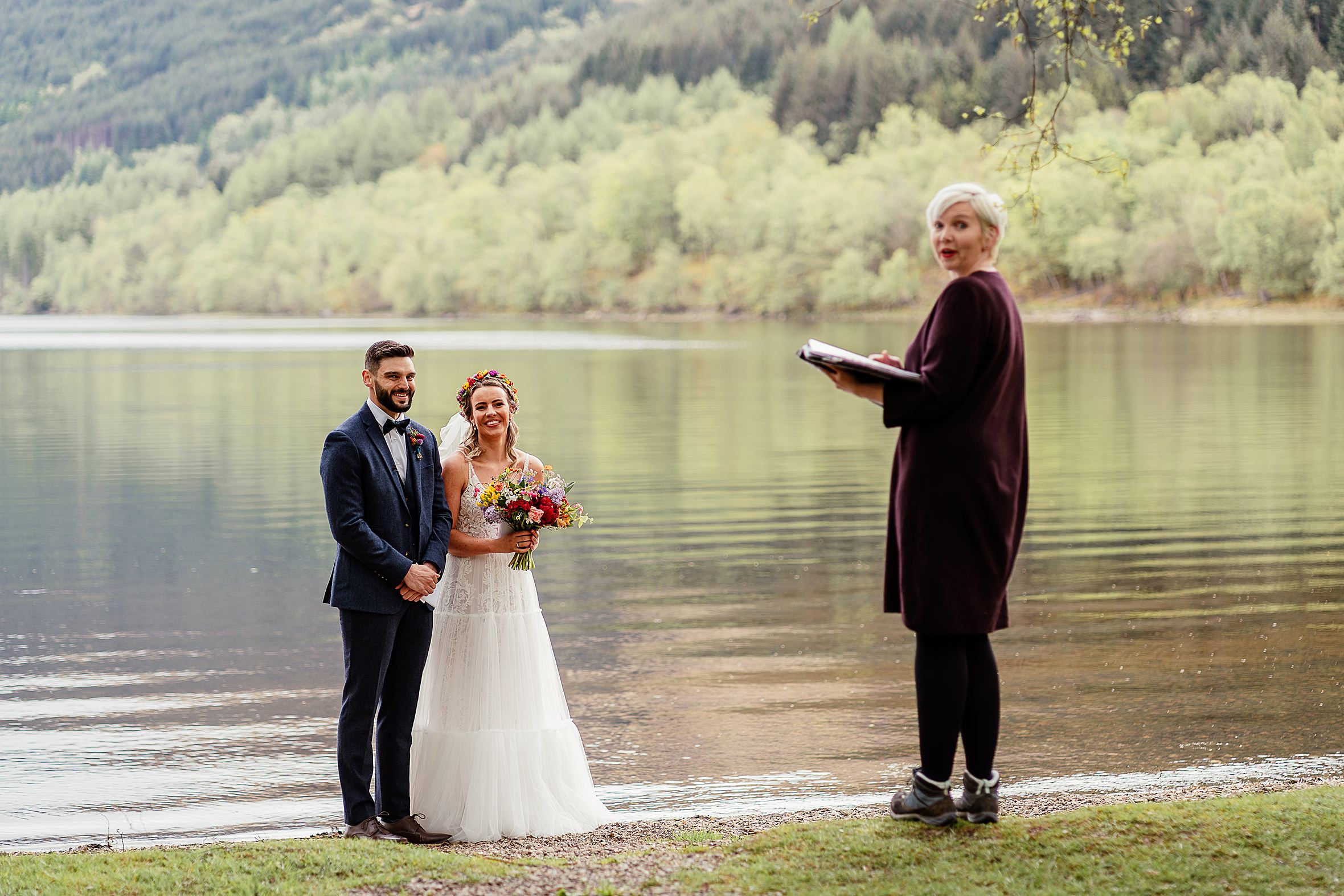 claire the humanist looking at camera on shore of loch with smiling bride and groom behind her green foresty hills in background humanist weddings scotland