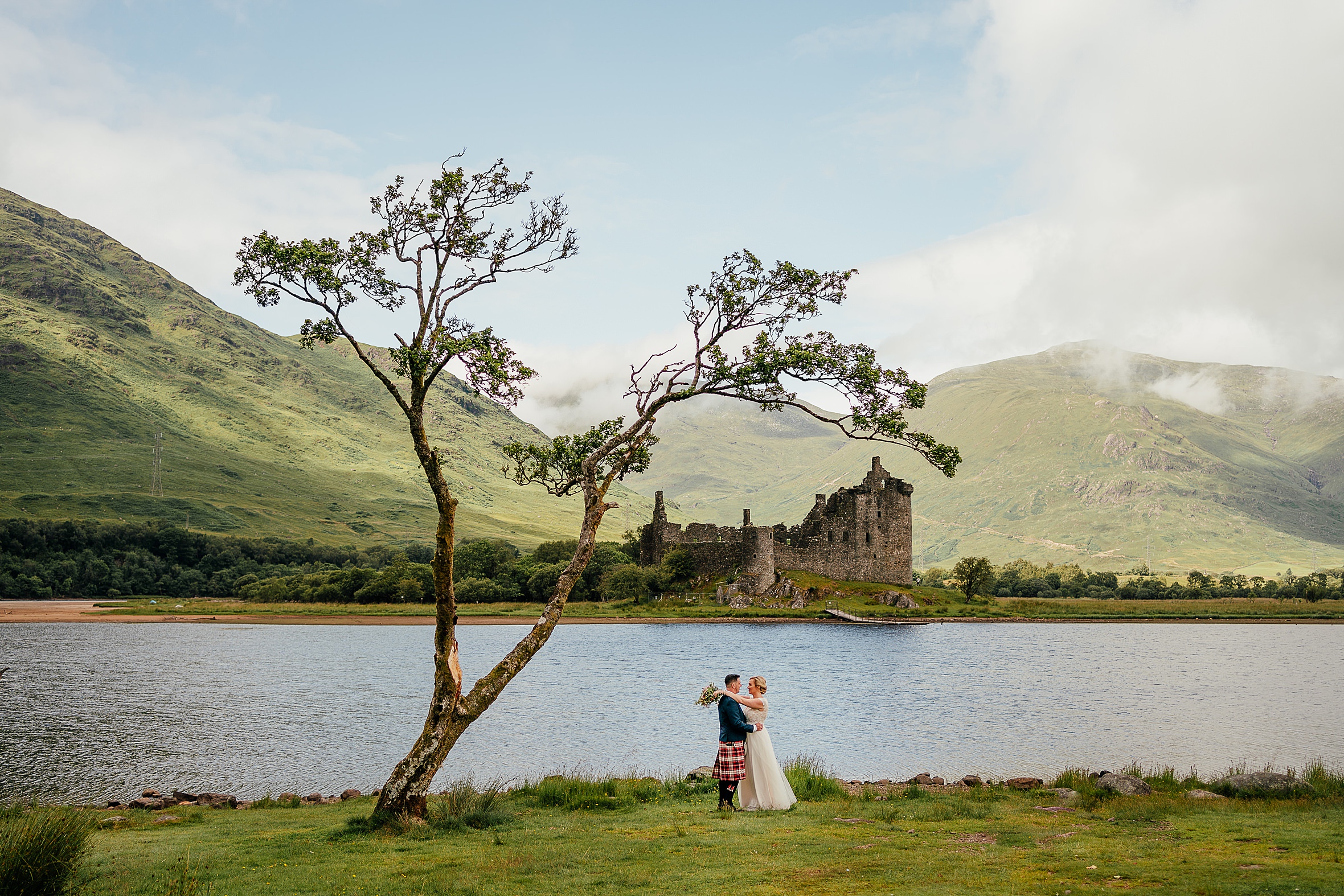 bride and groom holding each other on the shore of the loch next to a tree green mountains blue skies and castle in background kilchurn castle wedding