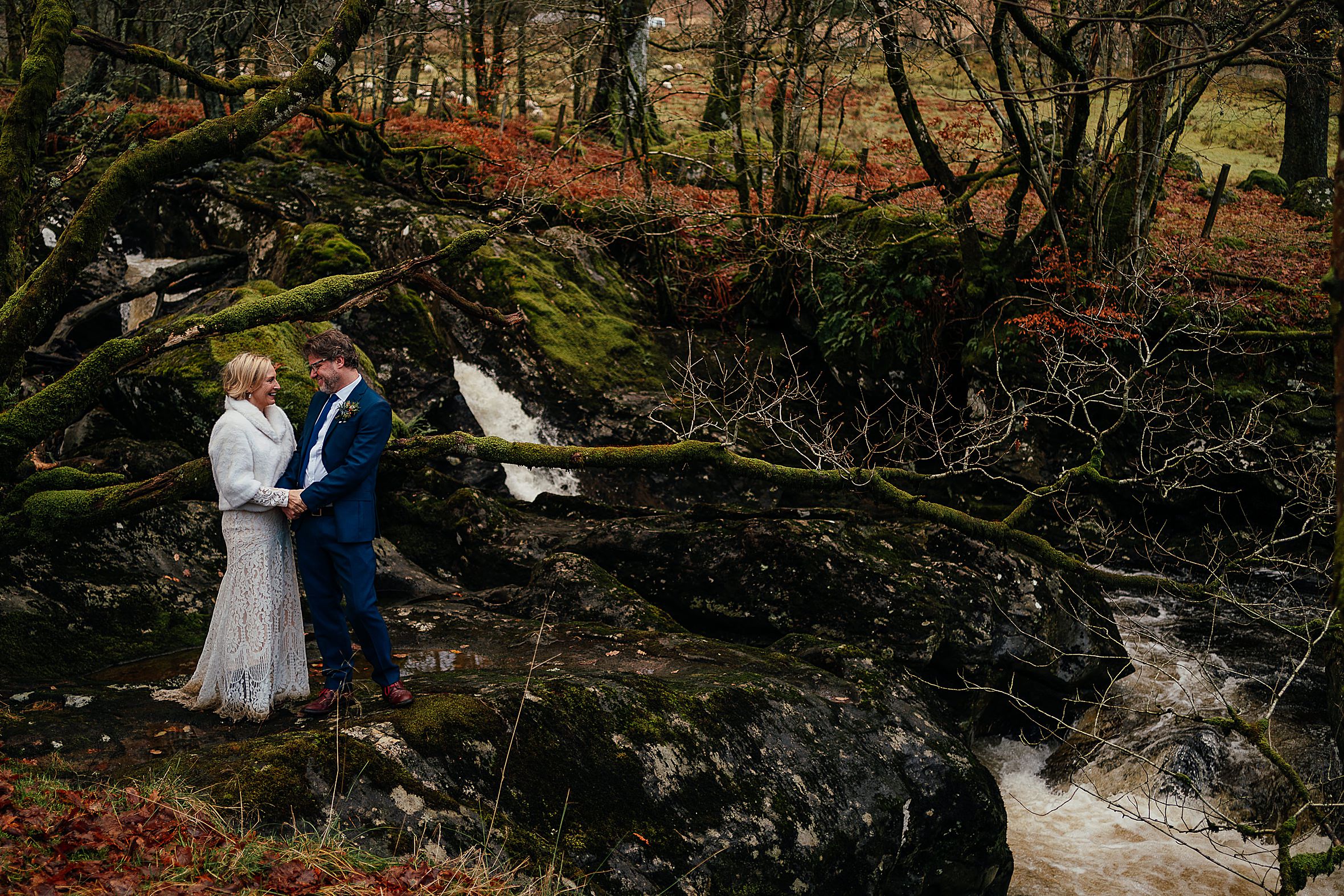 bride and groom holding hands and facing each other as they stand in woodland on rocks next to river scottish winter elopement